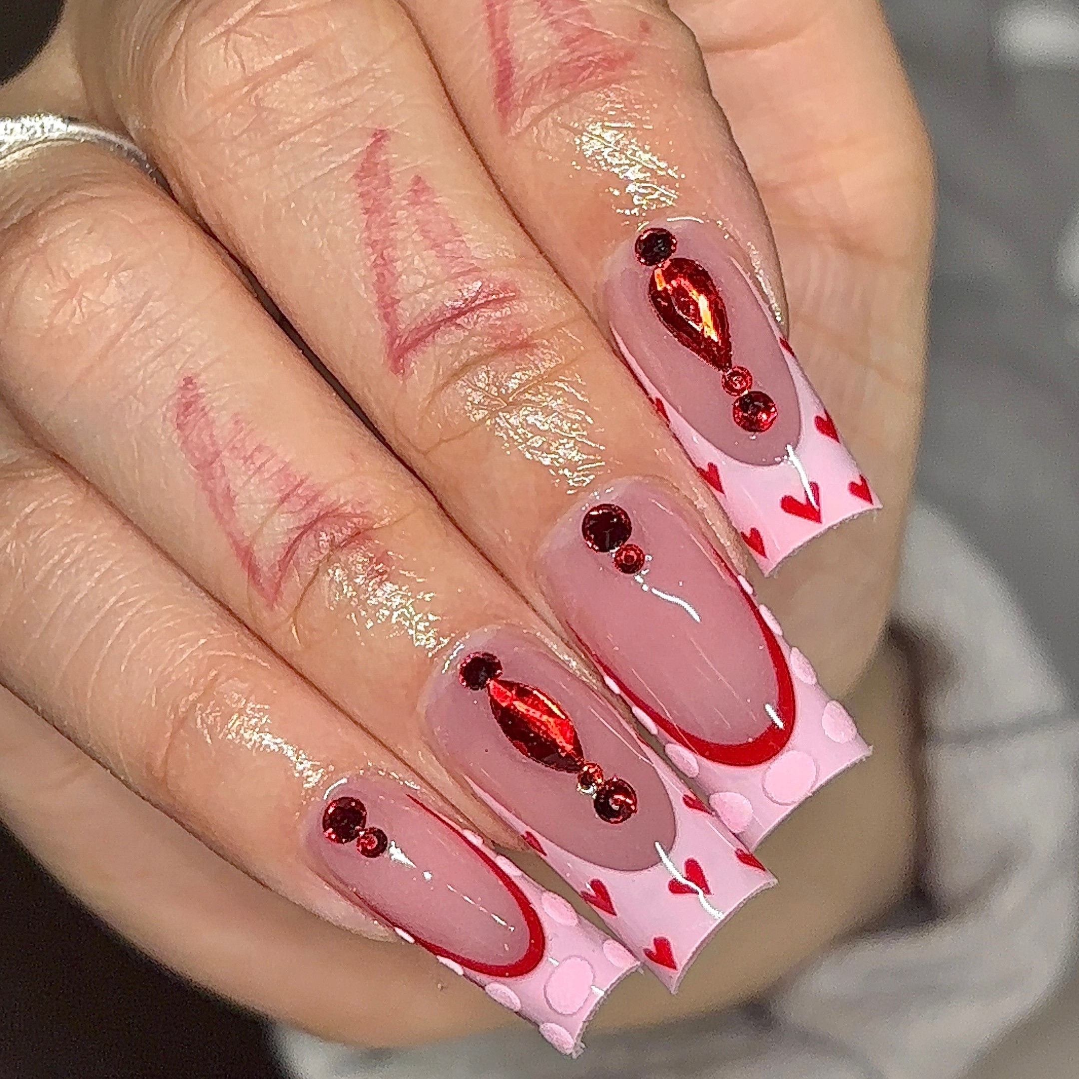 Medium square Gel-X nails with a nude pink base, red chrome heart accents, rhinestones, and Valentine's-inspired nail art by n4ilsbybellaa